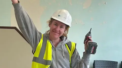 A woman wearing a white hard hat and a green/yellow high-vis waistcoat over a grey hoodie, holds a drill in her left hand and a hammer in her outstretched left hand. She stands in a building with green, peeling paint behind her and chairs stacked behind her.