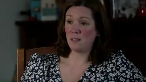 Nichola is pictured sitting at a table wearing a dark floral-patterned top, with a blurred home setting in the background.