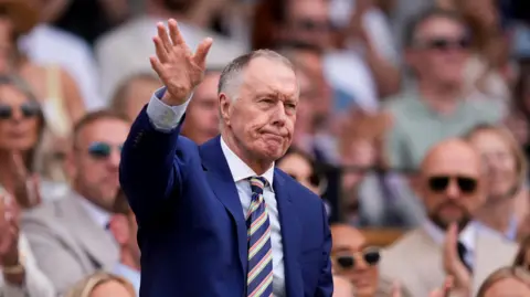 PA Media Sir Geoff Hurst is wearing a dark blue suit and a white shirt along with a striped tie and is waving. He is standing in a crowd of people, most of whom are wearing sunglasses.