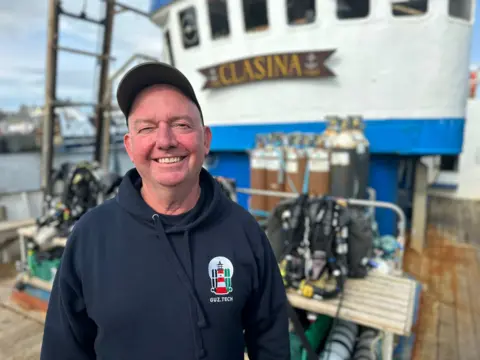 Diver Steve Mortimer smiles on board the Clasina vessel which took divers out to find the wreckage. We can see oxygen tanks and diving gear behind him