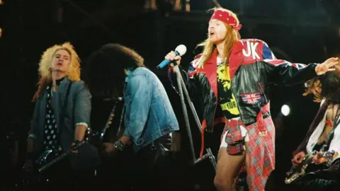 Getty Images Axl Rose singing onstage wearing a bandana and a leather jacket. Two guitarists and a bassist are visible in the background. 