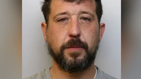 Mugshot of Atkinson. He has dark hair and a dark beard and is wearing custody uniform.