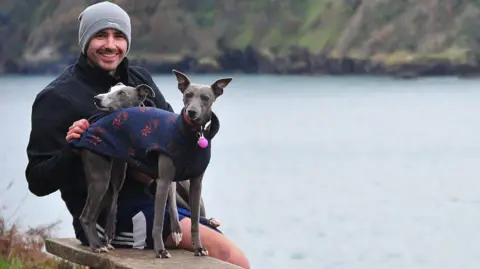 Shaun Reynolds A man wearing a grey hat, black shorts and a black coat, sitting on a bench with two grey whippets and smiling at the camera. There's a lake in soft focus in the background.