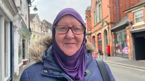 Zeinab looking into the camera and smiling; she's wearing a navy blue winter coat with a fluffy hood, and a purple hijab. She's stood on the pavement, with Wem's high street stretching out behind her, a combination of red brick buildings and white painted shop fronts. The sky is grey and cloudy.