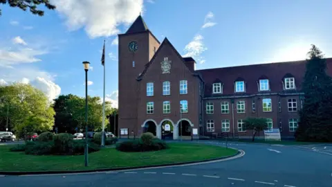 The red/brown brick headquarters of Spelthorne Borough Council, a building with slanted rooves, a clock tower and a green space and roads in front of it. There is a flagpole on the green space.