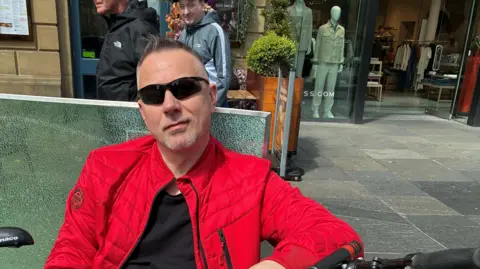 Lucas Krajewski sitting on a bench in Newcastle. He is wearing sunglasses and a red jacket. His bike is leaning beside him.