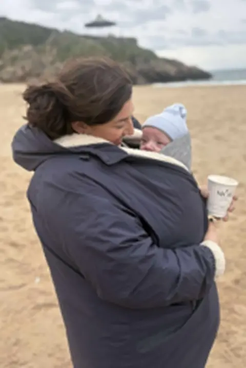 Ellie Atkins-Tate A woman on a beach in a blue coat. She has a baby tucked inside her coat and holds a coffee cup. She is on the beach.