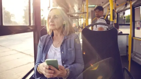 Getty Images A woman wearing a purple cardigan is listening to her phone through earplugs on a bus.  A man in a cap is looking in her direction.