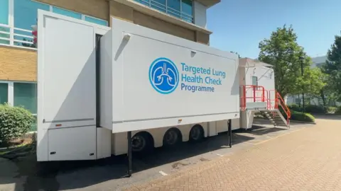 A large white truck trailer is parked in a car park. Targeted Lung Health Check Programme is written on the side in blue, along with a logo containing a graphic depicting a pair of lungs.