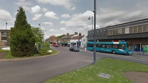 Google Roundabout with shrub-covered island. There is a blue bus which has stopped outside a Tesco Metro shop. There are other shops to the left, and cars on the roundabout. There are some clouds in the sky above. There is a grassed area in the foreground.
