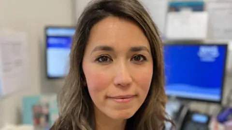 Dr Annie Lomas, who has long broan hair, looking directly at the camera in a GP surgery. In the background there are two computer screens, a phone and information n the wall. 