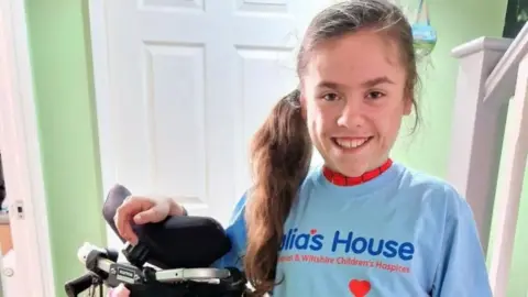 Julia's House Carmela, from Wareham wearing a light blue julia's house t-shirt over her superhero costume and stood next to her wheelchair