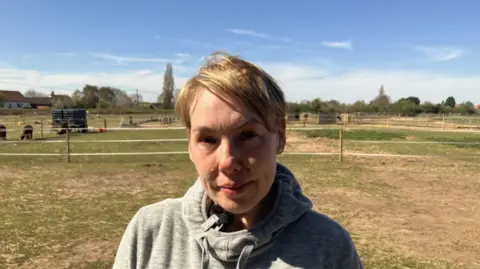 Lara King/BBC Julia Stewart - a woman with short blonde hair wearing a grey hoodie. She is standing in front of fencing with animals behind her.