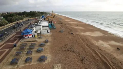 Southsea Coastal Scheme Beach with sea to the right and road and paths to the left. There are a number of huts and tables and chairs running alongside the rear of the sand.
