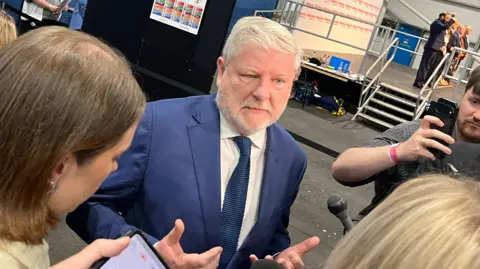 Angus Robertson, who has grey hair and a grey beard, in a blue suit in a counting centre 