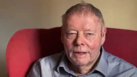 BBC/Genevieve Tudor A man with short dark hair and a blue-white checked shirt sits in a red armchair in front of a white wall.