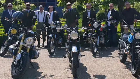 Paul Eaton A group of about a dozen men dressed dapper in ties or dinner jackets, some with arms around others, smiling to camera behind their classic motorbikes in the sunshine