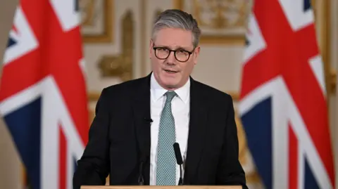 Sir Keir Starmer standing at a podium with two black microphones and two large Union Jack flags behind him