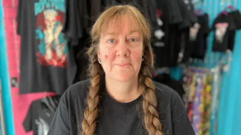 A middle aged woman with two long, light brown plaits in her hair and a fringe smiles. She is wearing a black top and has ear stretchers. She is stood in her alternative fashion shop and black graphic T-shirts can be seen behind her.