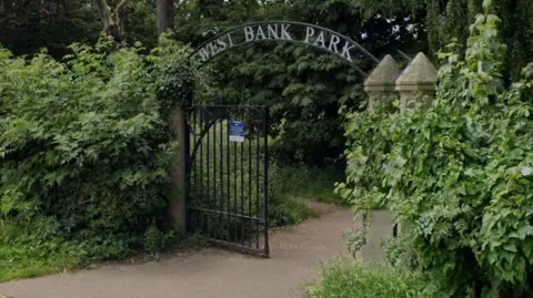 Black park gates with overgrown bushes and grass. A sign above the park gate reads West Bank Park.