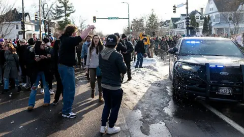 AFP via Getty Images A group of protesters stand in the middle of a road next to a parked sheriff's car. One woman can be seen shouting, while another person is pointing at the car. Snow is falling. 