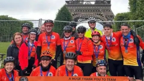 Deb Gascoyne A group of 16 people, all wearing orange lycra tops, with the Eifell Tower in the background. Many are wearing cycle helmets and they are holding up a large placard promoting their London-Paris bike ride.