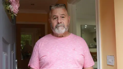 Paul Thomas wearing a pink t-shirt is standing in his doorway and looking straight at the camera. 