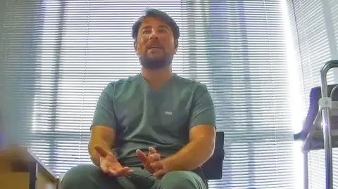 BBC Ricky Sawyer, filmed undercover in his small pop-up clinic at a London office, sits next to a metal trolley wearing green uniform. He is a man in his 20s with straight brown hair, a moustache and beard.