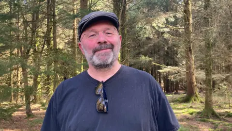 Julie Mariotti/BBC A man in a black T-shirt smiles at the camera. He's standing in the woods on a sunny day.