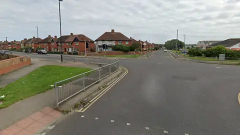 The four-way junction has red-bricked semi-detached houses on the corners, and an Iceland store on the right. There are grey fences and yellow lines on each of the corners, and a crossing area on each side, but further down.