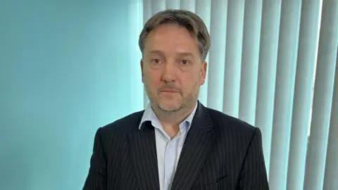 A man with a curtain style haircut in a black blazer and blue shirt.