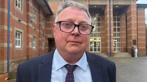 A man with short grey hair and dark framed glasses in a blue suit and blue tie outside a brown brick building with large windows