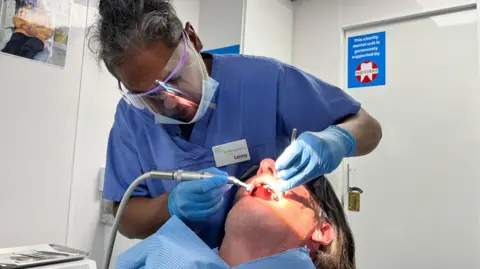 Martin Giles/BBC A dentist in blue scrubs and a face visor peers into a patient's mouth as he uses a piece of electrical equipment on their teeth. The dentist has a name badge: Lenny.