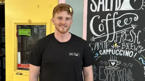 A man in a black t-shirt with a logo and a cafe sign behind him and a yellow door.