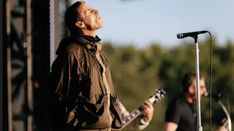 Liam Gallagher has his eyes closed with his head facing the sky while on stage. He is wearing a brown parka coat. To his left the arm of a band member playing guitar and Noel Gallagher playing guitar and singing, both blurred, can also be seen.