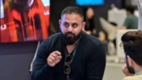 LinkedIn Fasial Tariq has a beard and moustache, he wears a blue jumper with sunglasses tucked into the V-neck. 