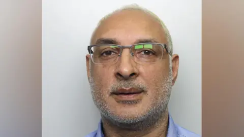 A man in his 50s, pictured in a police mugshot. He has very thin white hair, white stubble and glasses. He is wearing a blue buttoned shirt and staring directly at the camera.