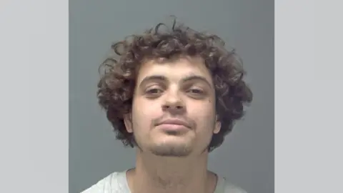 A mugshot of Logan Burnett, who is smiling at the camera with his head tilted upwards. He has dark, curly hair and brown eyes. He has a small amount of facial hair.
