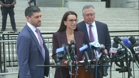 Luigi's lawyer wearing a brown suit, speaks into mic.