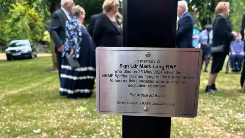 The plaque reads "In Memory of Sqn Ldr Mark Long RAF who died on 25th May 2024 when his BBMF Spitfire crashed flying to this memorial site to honour this Lancaster crew during the dedication ceremony. Per Ardua ad Astra. Kindly funded by RAFA Cranwell Branch."