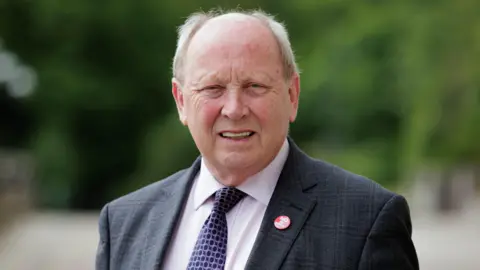 PA Media Jim Allister stands outside, wearing a grey suit white shirt and purple patterned tie. He has grey hair.