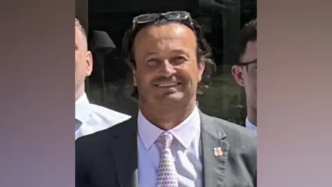 A man is smiling and wearing a suit jacket over a white shirt, with a patterned tie. He has propped his glasses on his forehead.