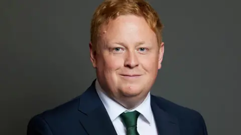 Christopher Bloore/House of Commons/Laurie Noble A man with ginger hair wearing a navy suit jacket, white shirt and green tie smiles at the camera