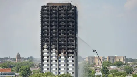 Burnt-out Grenfell tower block. Firefighters on a turntable ladder are dousing the building with water. There are other flats and rooftops in the distance.