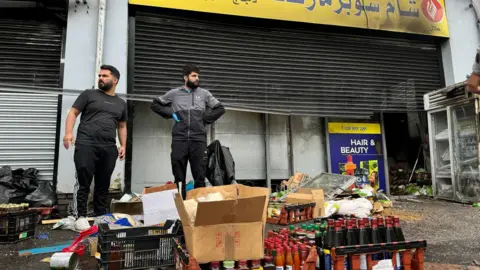 Pacemaker Two shopkeepers outside fire-damaged store on Donegall Road with damaged stock strewn across the pavement