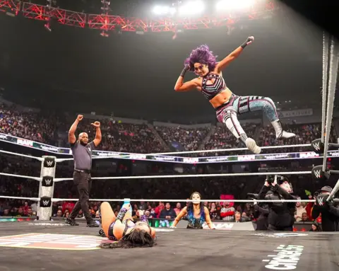 Getty Images Bayley leaps from the top turnbuckle in a wrestling ring, delivering an elbow drop to a prone opponent.