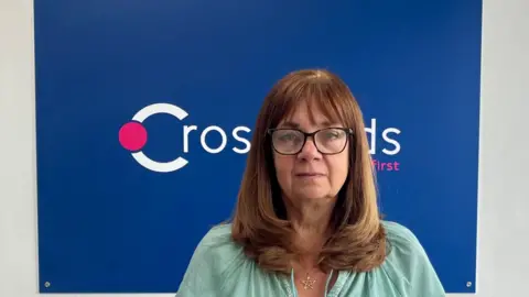 BBC Jackie Betteridge, a woman with brown hair and a fringe, wears glasses. She is photographed in front of a predominantly blue sign that reads Crossroads.