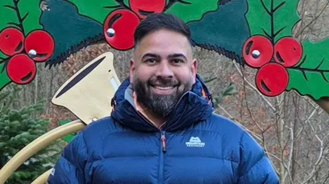 Handout Maj Mahmood with black hair and beard wearing a navy blue puffer jacket standing in a woodland in front of a decorative holly and bell. He is smiling. 
