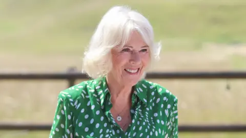 Queen Camilla is wearing a green and white polka dot blouse. She is standing outside. A grass field and wooden fence is in the background.