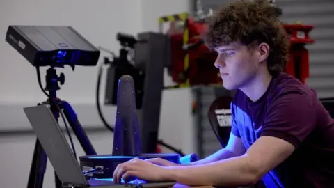 Harper Adams University A student with curly brown hair stares at a laptop in some kind of room with equipment behind him. The light from the laptop illuminates his mauve T-shirt.